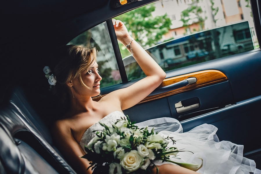 Portrait of a beautiful blonde bride with bouquet sitting in  wedding car and looking at somebody or something through the window. Wedding day. Daylight.